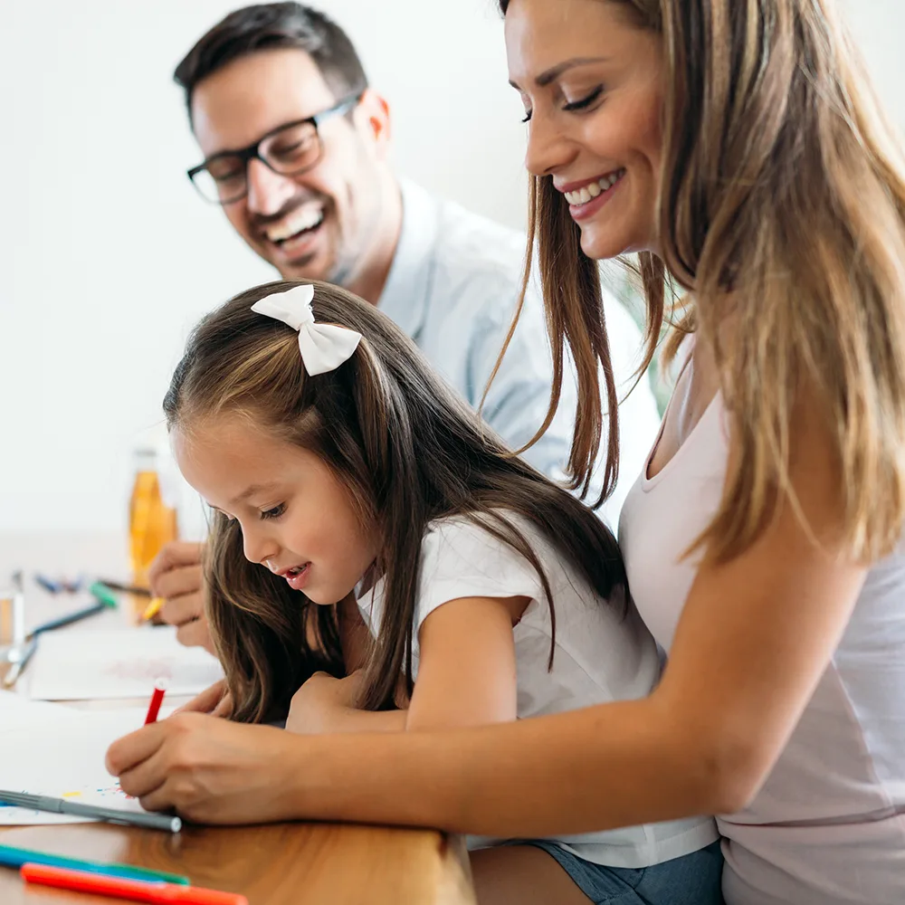 A laughing mother and father helping their child write in a book at a Red Balloon Child Care Center daycare
