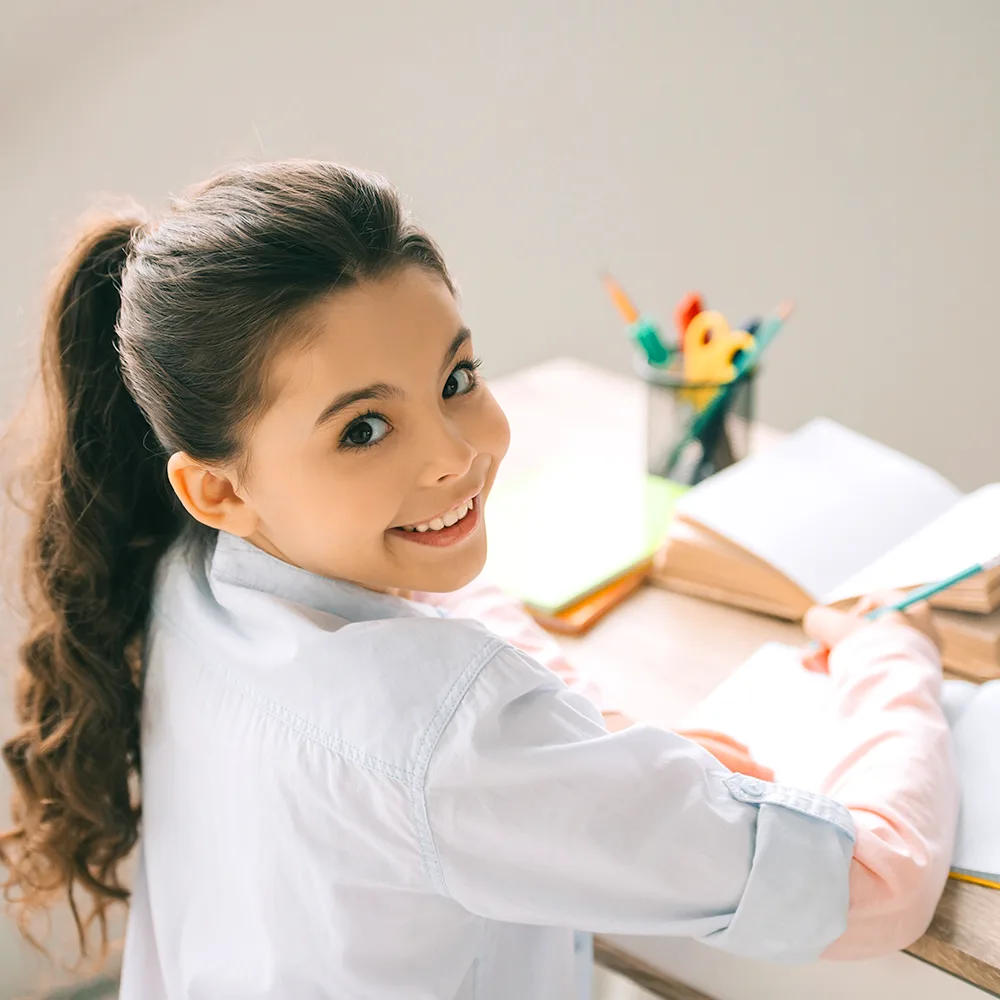 A child at a desk, turning and smiling, at a Red Balloon Child Care Center daycare.