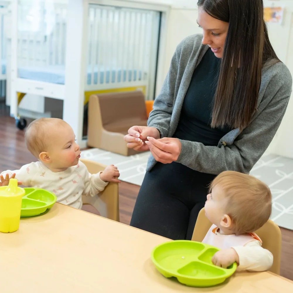 A teacher feeding two small toddlers at a table at a Red Balloon Child Care Center daycare